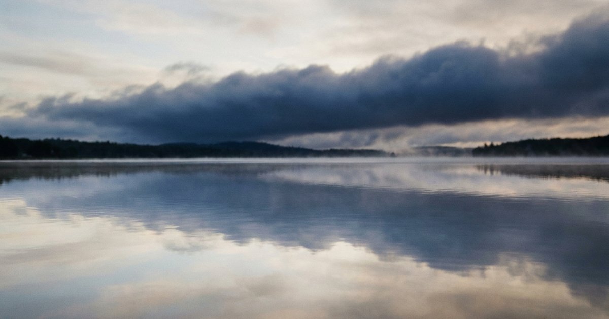 Un lago all’alba con l’acqua in primo piano immobile come uno specchio e una fascia di nubi scure sulla riva opposta