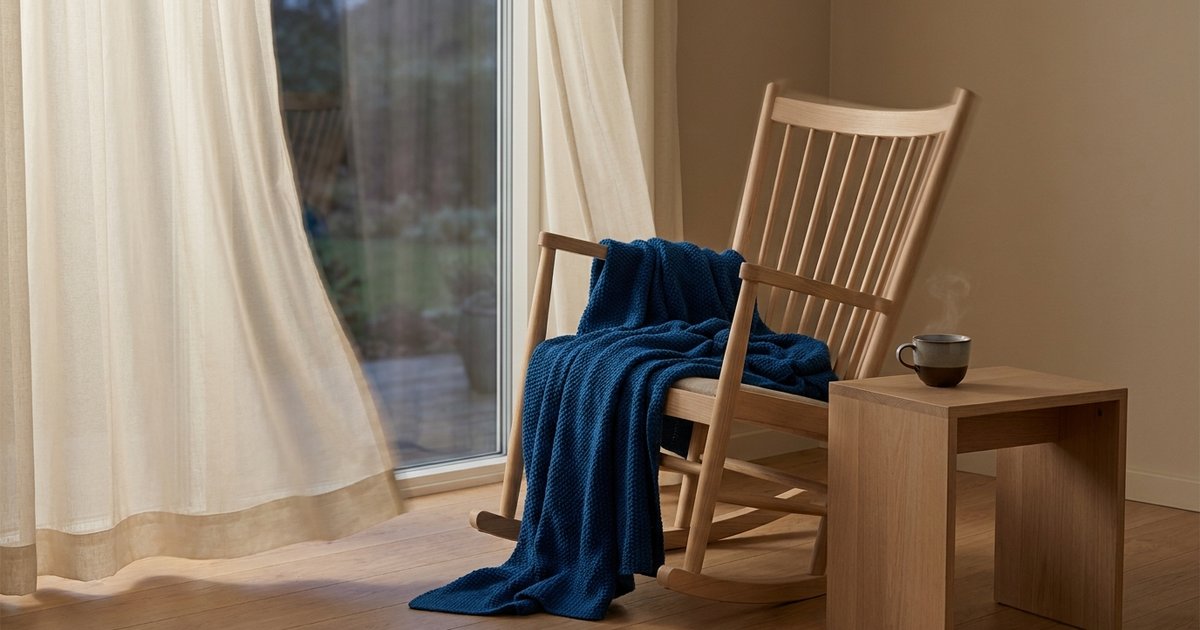 Irrequietezza: una sedia a dondolo in legno chiaro con una coperta blu, accanto a una tazza fumante su un tavolino e a una tenda leggera mossa dal vento vicino a una porta finestra